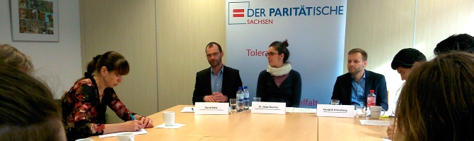 Auf einer Pressekonferenz beim Paritätischen Wohlfahrtsverband Sachsen sitzen Menschen an einem Tisch. Vor drei Personen stehen Namenschilder und im Hintergrund ist das Logo des Paritätischen (ein rotes Gleichheitszeichen in einem blauen Rahmen) zu sehen.