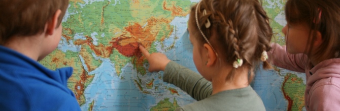 Zwei Mädchen und ein Junge im Kindergartenalter stehen vor einer Weltkarte. Das braunharige Mädchen mit Zöpfen in der Mitte zeigt mit dem Finger den Himalaya auf der Weltkarte.