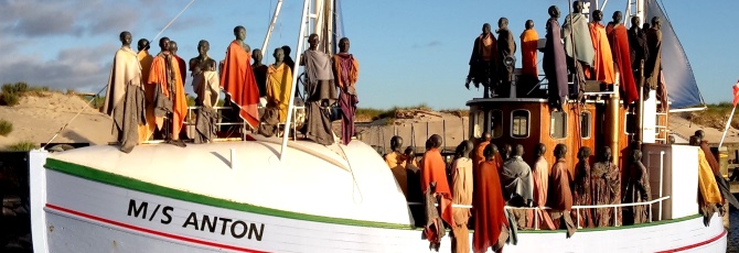 Auf einem weißen Holzschiff stehen lebensgroße Figuren mit dunkler Haut und in langen bunten Gewändern. Die Holzfiguren erwecken den Eindruck, dass man auf ein mit Flüchtlingen voll besetztes Boot blickt.