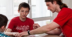 Ein junger Mann mit langen braunen Haaren und rotem T-Shirt sitzt mit zwei Jungs an einem Tisch und spielt ein Brettspiel.