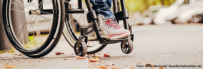 Die Räder eines Rollstuhls stehen am Bordstein einer Straße.