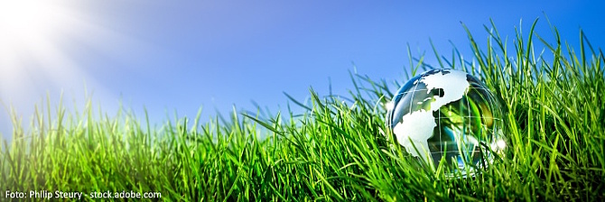 Symbolbild: Eine gläserne Weltkugel liegt in einer grünen Wiese auf die aus einem blauen Himmel die Sonne scheint.