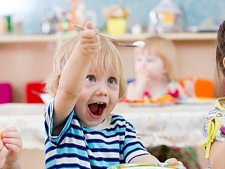 Drei kleine Kinder sitzen in der Kita am Tisch und essen. Das Mittlere Kind hebt freudig seinen Löffel in die Höhe. (Foto: Andrey Kuzmin/ fotolia.com)