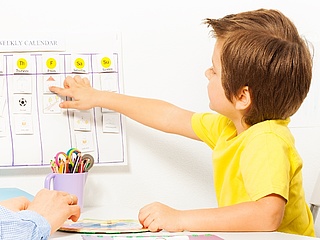 Ein Junge sitzt an einem Tisch und zeigt auf einen Wochenplan an der Wand. (Foto: Sergey Novikov/ Fotolia.com)