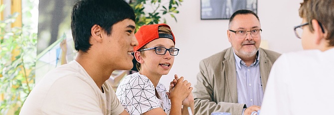 Drei Jugendliche und ein Mann sitzen an einem Tisch und Diksutieren. Das Bild illustriert einen Artikel über Mitbestimmung im SOS-Kinderdorf Zwickau des Magazins anspiel. (Foto: Michael Bader, Archiv SOS-KInderdorf e.V.)