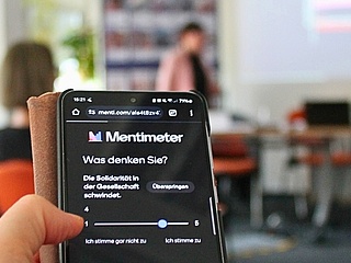 Im Vordergrund ein Smartphone-Bildschirm mit einer digitalen Abstimmung. Im unscharfen Hintergrund blicken Menschen in einem Seminarraum auf die an die Wand projizierten Abstimmungsergebnisse.