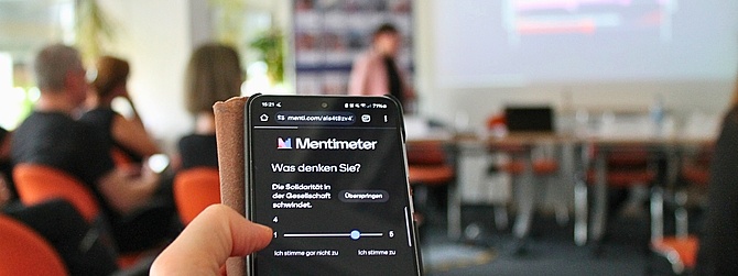 Im Vordergrund ein Smartphone-Bildschirm mit einer digitalen Abstimmung. Im unscharfen Hintergrund blicken Menschen in einem Seminarraum auf die an die Wand projizierten Abstimmungsergebnisse.
