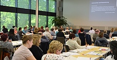 Rund 100 Personen sitzen in einem hellen und großen Konferenzsaal zu Gruppen an Tischen. Sie hören aufmerksam einer Frau mittleren Alters bei ihrem Vortrag zu. An der hohen Saalwand im Hintergrund ist eine Präsentation zu sehen.