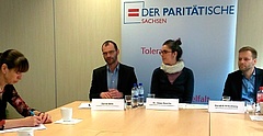 Auf einer Pressekonferenz beim Paritätischen Wohlfahrtsverband Sachsen sitzen Menschen an einem Tisch. Vor drei Personen stehen Namenschilder und im Hintergrund ist das Logo des Paritätischen (ein rotes Gleichheitszeichen in einem blauen Rahmen) zu sehen.