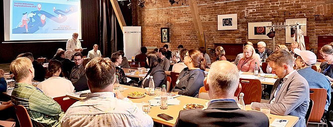 Menschen sitzen an Tischen auf der Regionalkonferenz in Leipzig. Von einem Podium spricht der Landesvorsitzende und informiert zum Zukunftskonzept Parität 2030.