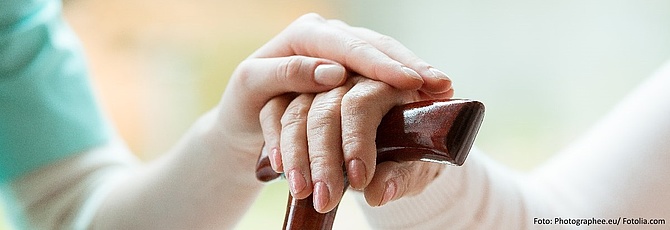 Auf der Hand eines älteren Menschen, die auf dem Griff eines Gehstocks liegt, liegt die Hand einer Pflegekraft.