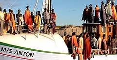 Auf einem weißen Holzschiff stehen lebensgroße Figuren mit dunkler Haut und in langen bunten Gewändern. Die Holzfiguren erwecken den Eindruck, dass man auf ein mit Flüchtlingen voll besetztes Boot blickt.