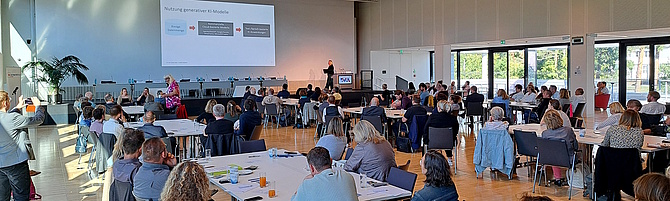 Mitgliederversammlung 2025: In einem hellen Saal sitzen Menschen an Tischen und hören einer Rednerin zu.